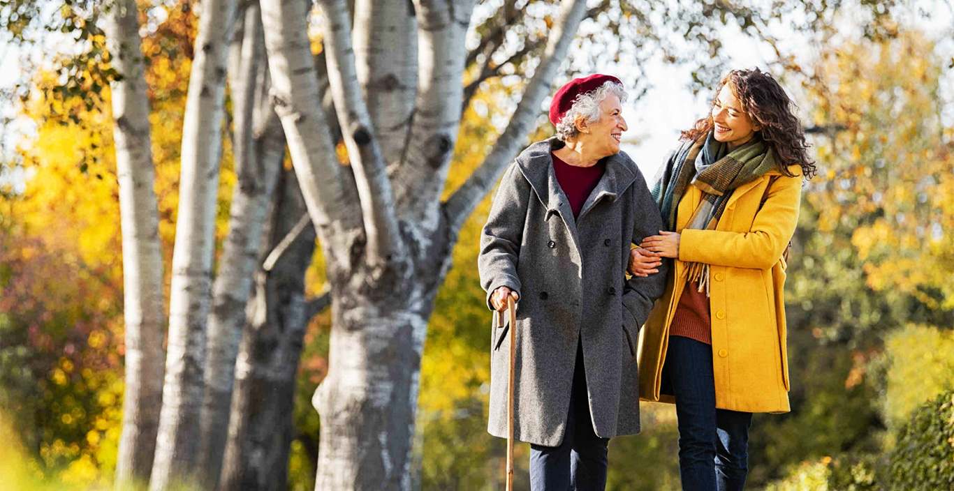 Support worker and older woman walking arm in arm through an autumn park